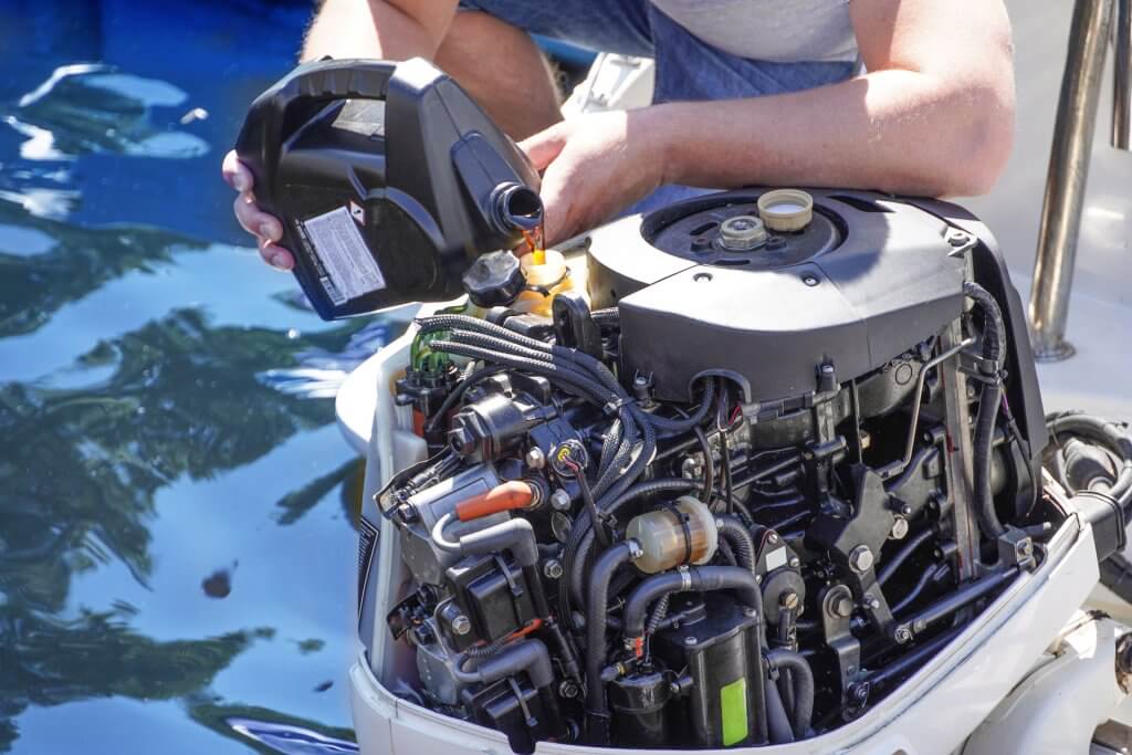 Ruke ulijevaju svježe ulje iz crne kanistre u otvoreni vanbrodski motor na krmi plovila, zamjena ulja uz more.